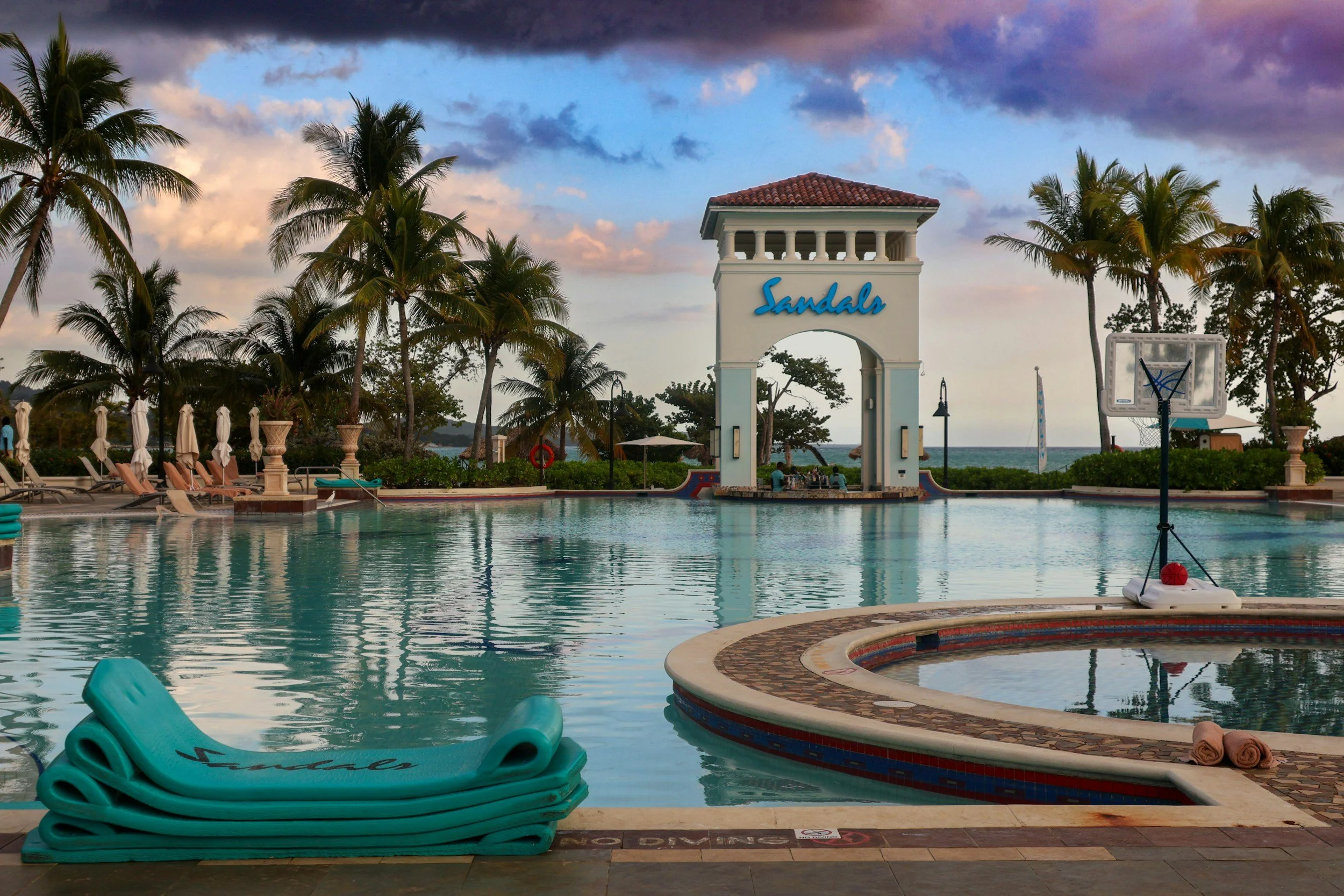 A swimming pool area at sunset with a sign that says 'Sandals'. The pool is surrounded by palm trees, lounge chairs with umbrellas, and a basketball hoop, with the ocean visible in the background.