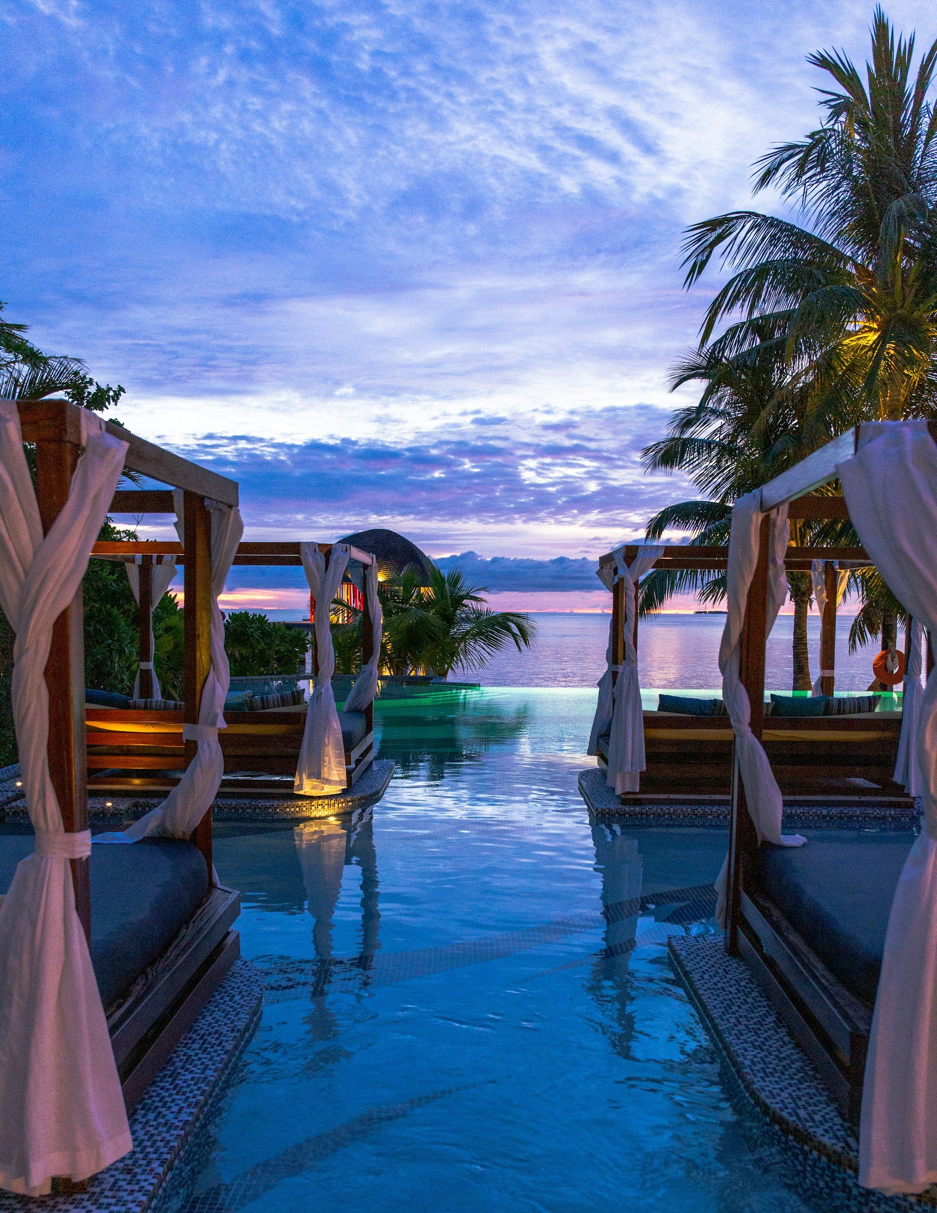 Overwater cabanas with draped curtains overlooking a swimming pool and ocean at sunset, surrounded by palm trees.