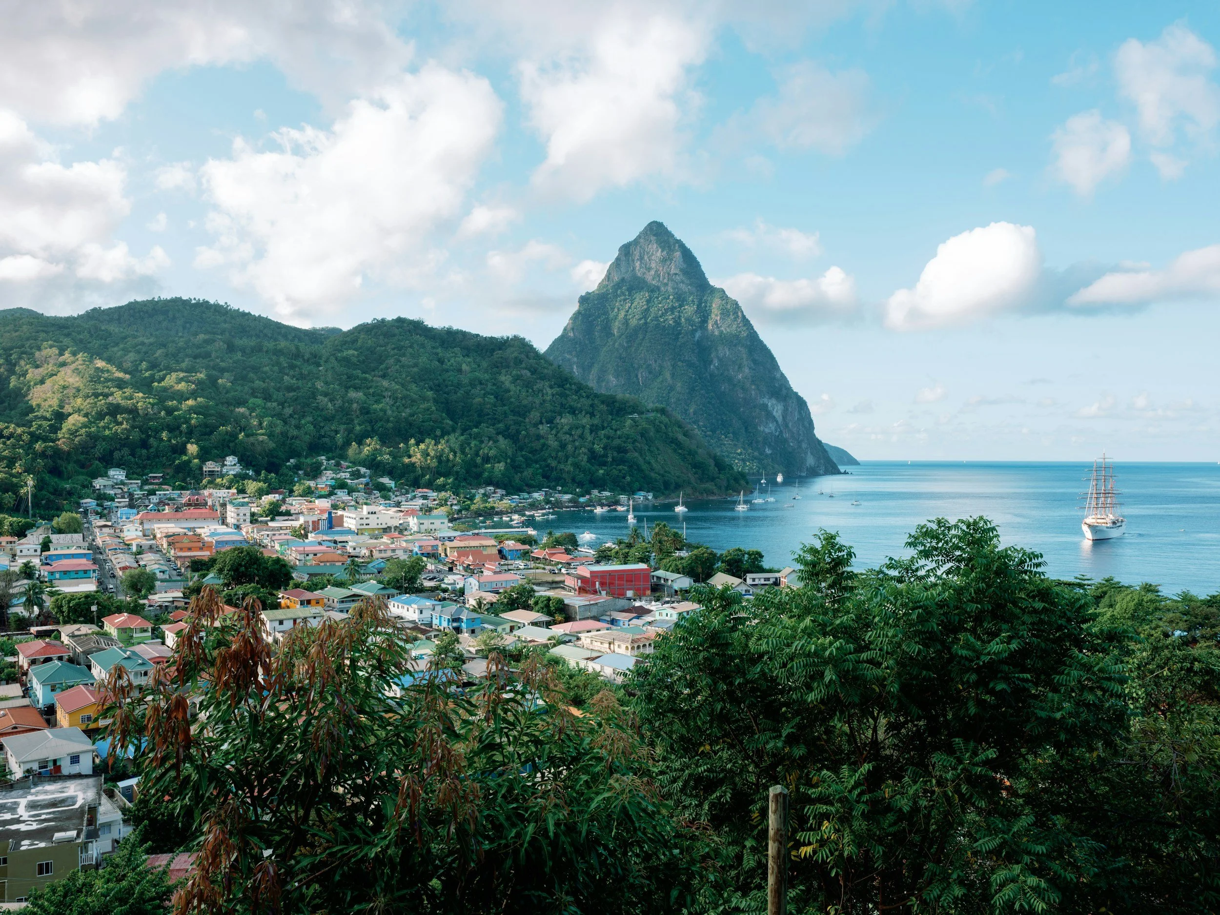 A coastal town with colorful houses at the base of a lush green mountain, featuring a prominent peaked mountain in the background. Several boats are anchored in the calm bay, and part of a sailing ship is visible on the right side of the image. The sky is partly cloudy with patches of blue.