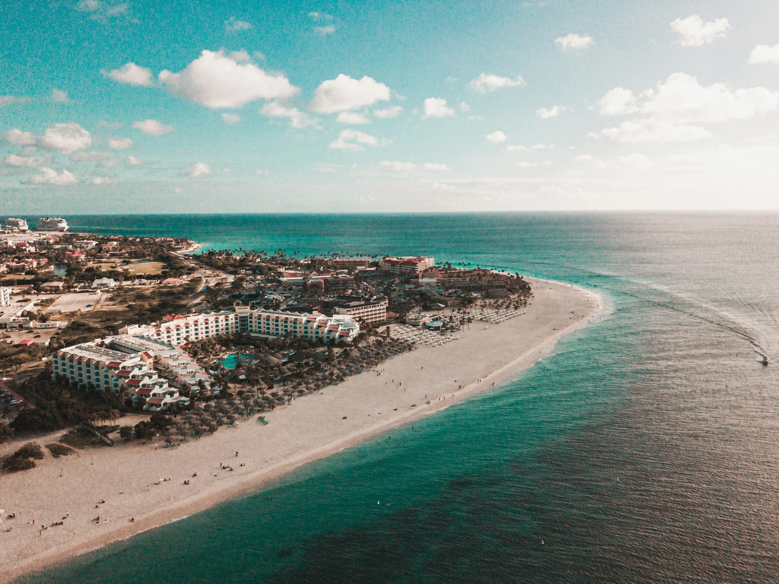 Aerial view of a coastal city with beaches, high-rise hotels, and palm trees along the shoreline, under a partly cloudy sky.