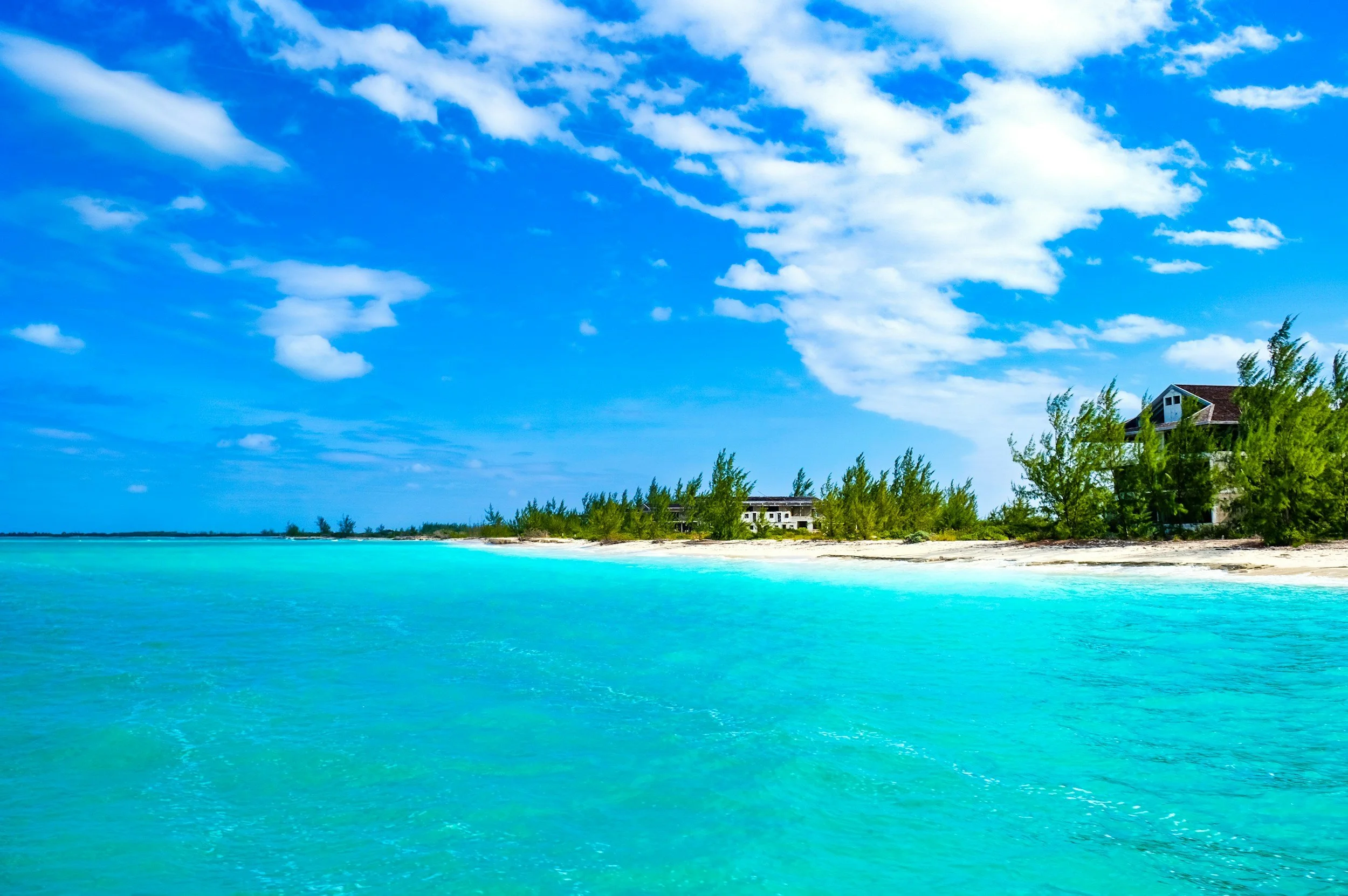 A tropical beach with turquoise waters, a sandy shoreline, green trees, and a house with a sloped roof under a blue sky with scattered clouds.