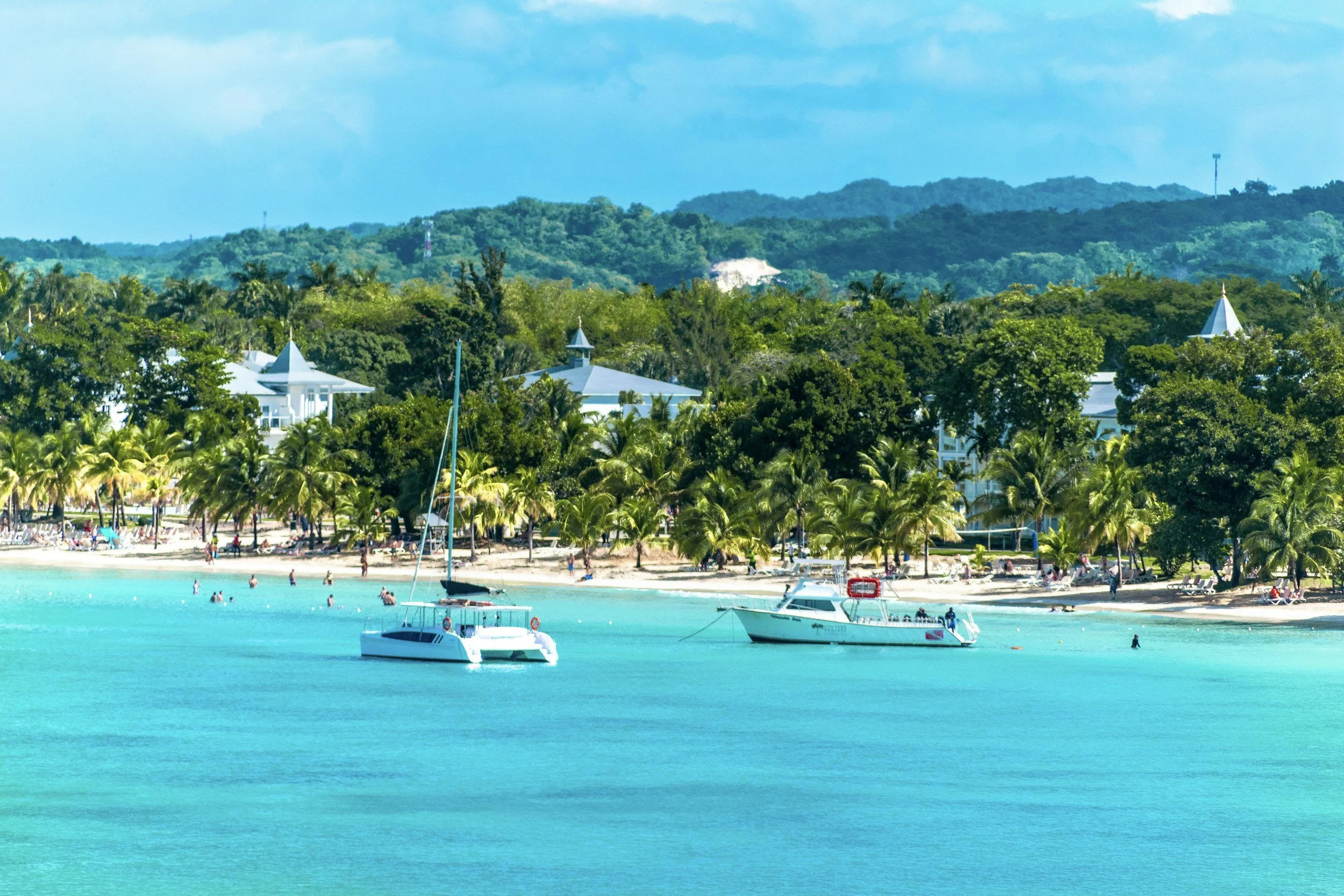 Sandy beach with palm trees, people swimming and sunbathing, and boats floating on turquoise water. Overlooking a lush green hillside with white buildings and a clear sky.