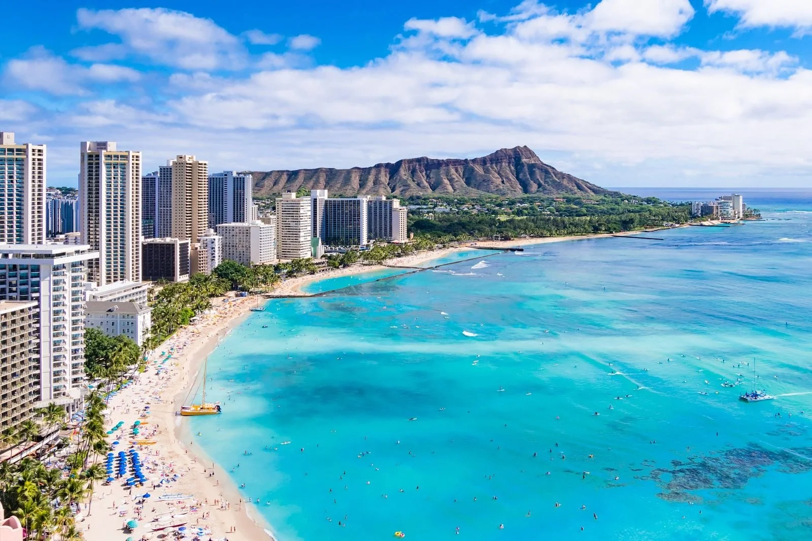 Aerial view of Waikiki Beach with high-rise hotels, sandy shore, turquoise water, and Diamond Head mountain in the background on a clear day.