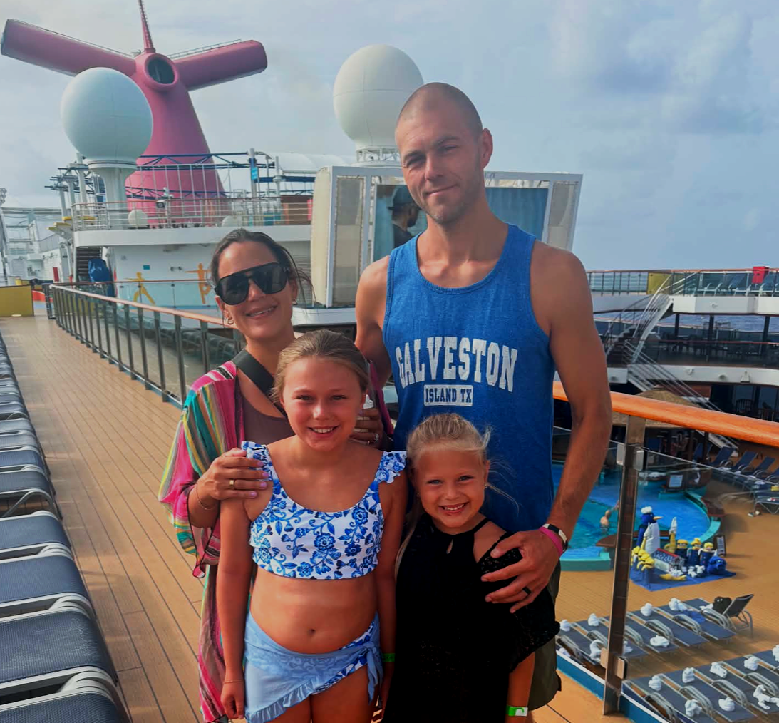A family of four on a cruise ship deck, smiling at the camera with the ship's amenities visible in the background.