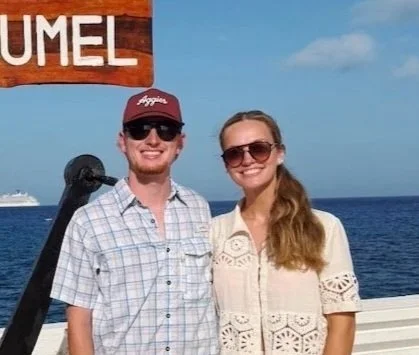 A smiling man and woman standing on a boat with the ocean in the background, wearing sunglasses and casual clothing.