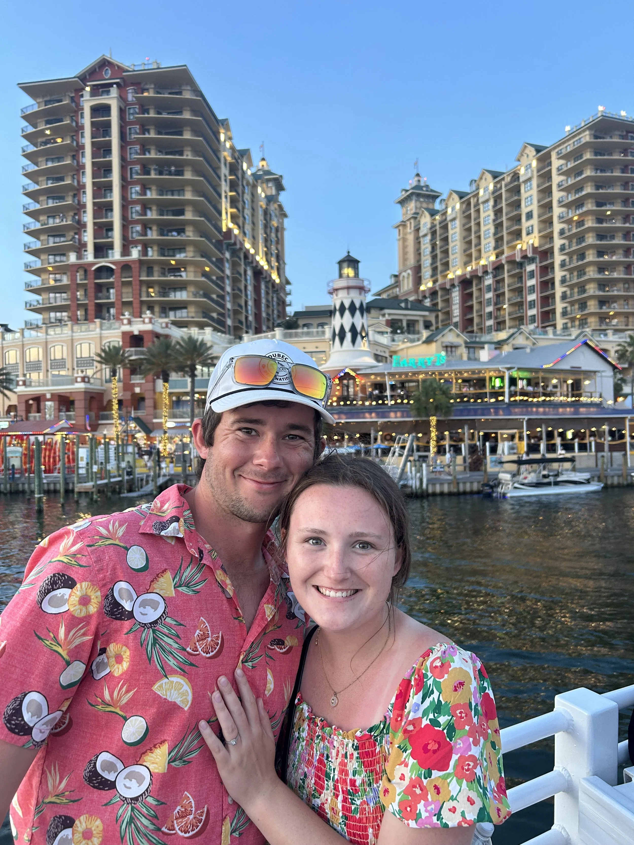 A young man and woman smiling together on a dock by the water, with tall colorful buildings and a lighthouse in the background, during daytime.
