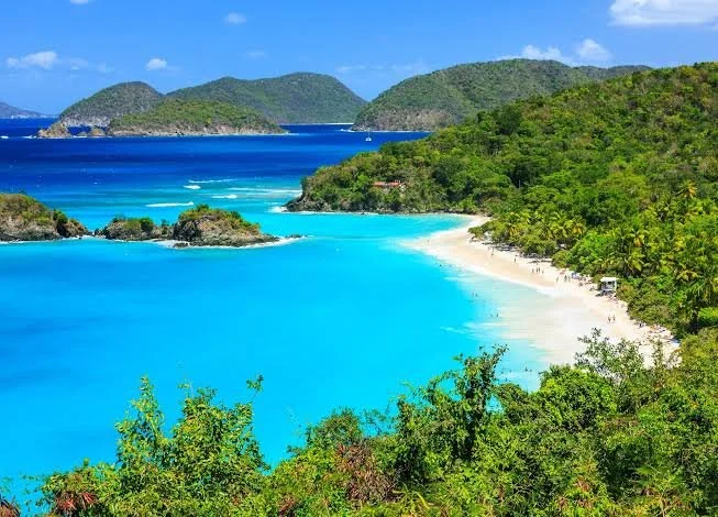 A tropical beach with turquoise water, white sand, lush green forest, and islands in the background.