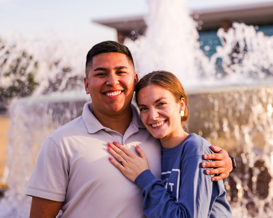 A smiling couple hugging outdoors in front of a fountain.