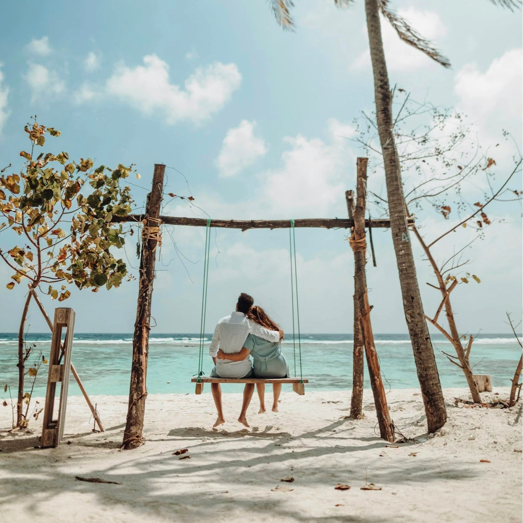 A couple sitting on a swing on a beach, with the ocean and sky in the background. The swing is framed by wooden and leafy structures.
