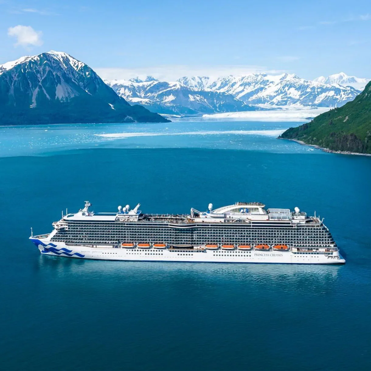 A large cruise ship sailing in icy blue water with snow-capped mountains and green forests in the background.