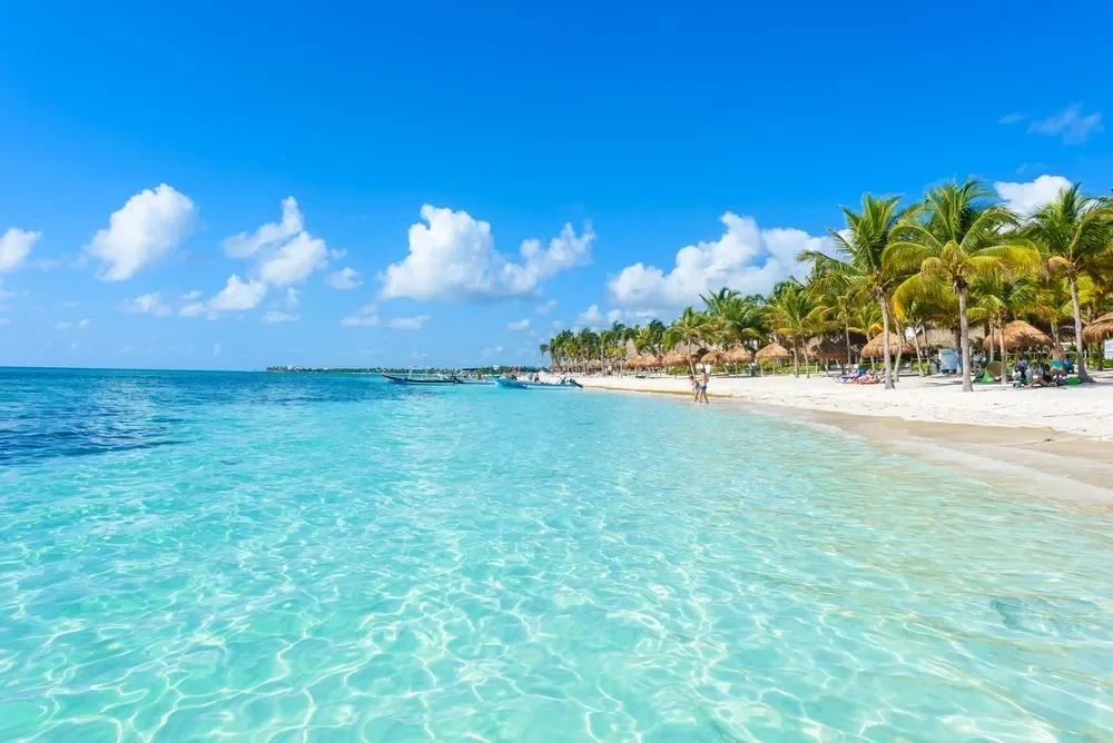 Tropical beach with clear blue water, white sandy shore, palm trees, and thatched-roof huts under a partly cloudy sky.