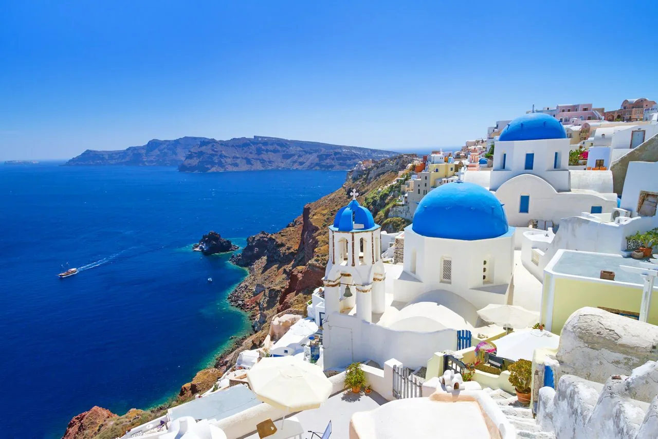 A scenic view of Santorini, Greece, featuring white buildings with blue domes on a hillside overlooking the ocean on a sunny day.