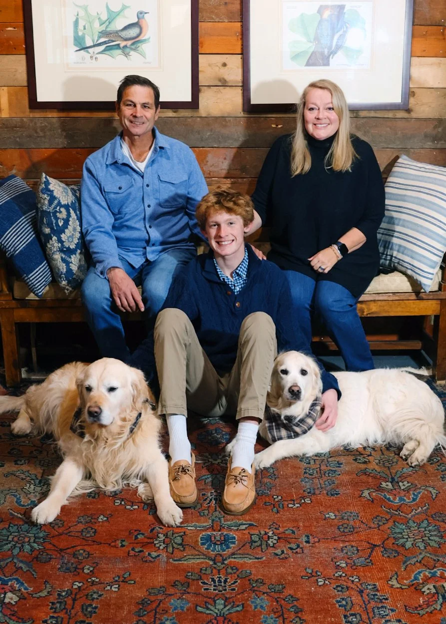 A family of three with two dogs sitting on a colorful patterned rug in a cozy, wood-paneled room with artwork of birds on the wall behind them.