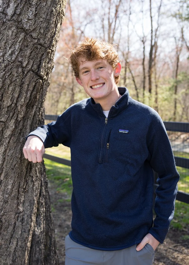 A young man with curly red hair and a big smile standing outside next to a tree, wearing a navy fleece jacket and gray pants, with a blurred background of trees and a black fence.