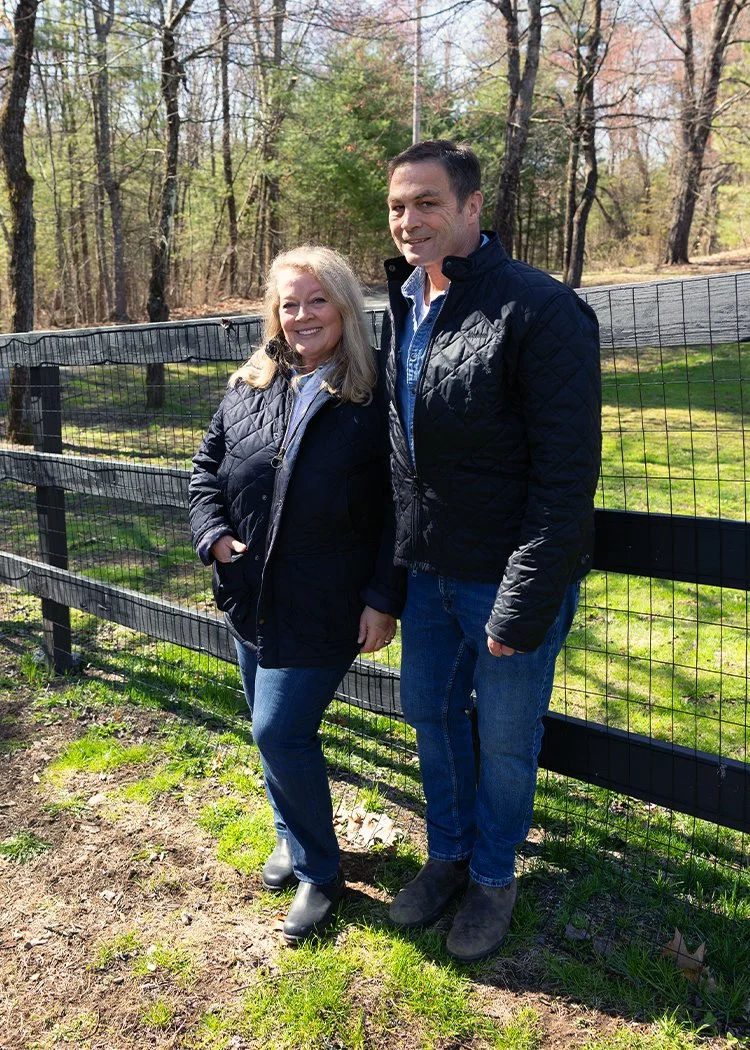 A smiling woman and man standing outdoors next to a wooden fence with trees in the background. They are both wearing dark jackets and jeans, with the woman in boots and the man in shoes.