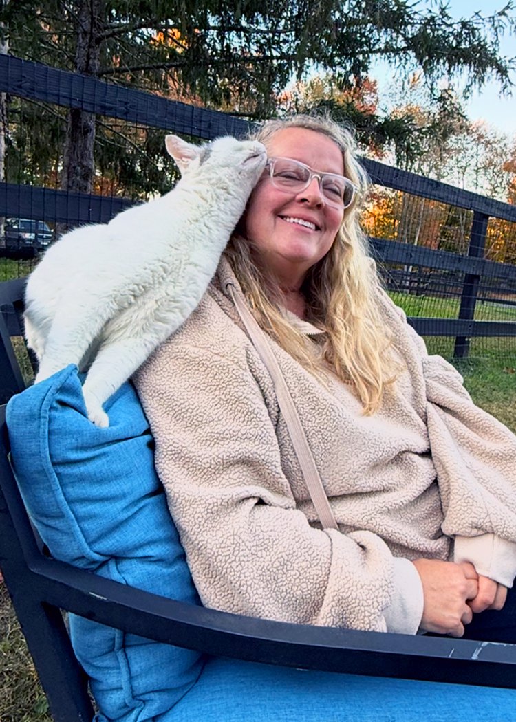 A woman smiling in outdoor setting with a white cat nuzzling her face, sitting on a blue cushion on a chair, with a fence and trees in the background.