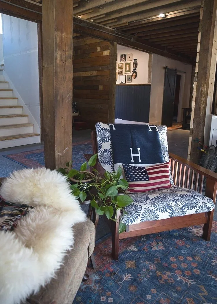 Cozy living room corner with a wooden armchair, decorative pillows featuring American flag and world map patterns, a plant on the armrest, a furry blanket or throw, rustic wood beams, stairs, and wall decor.