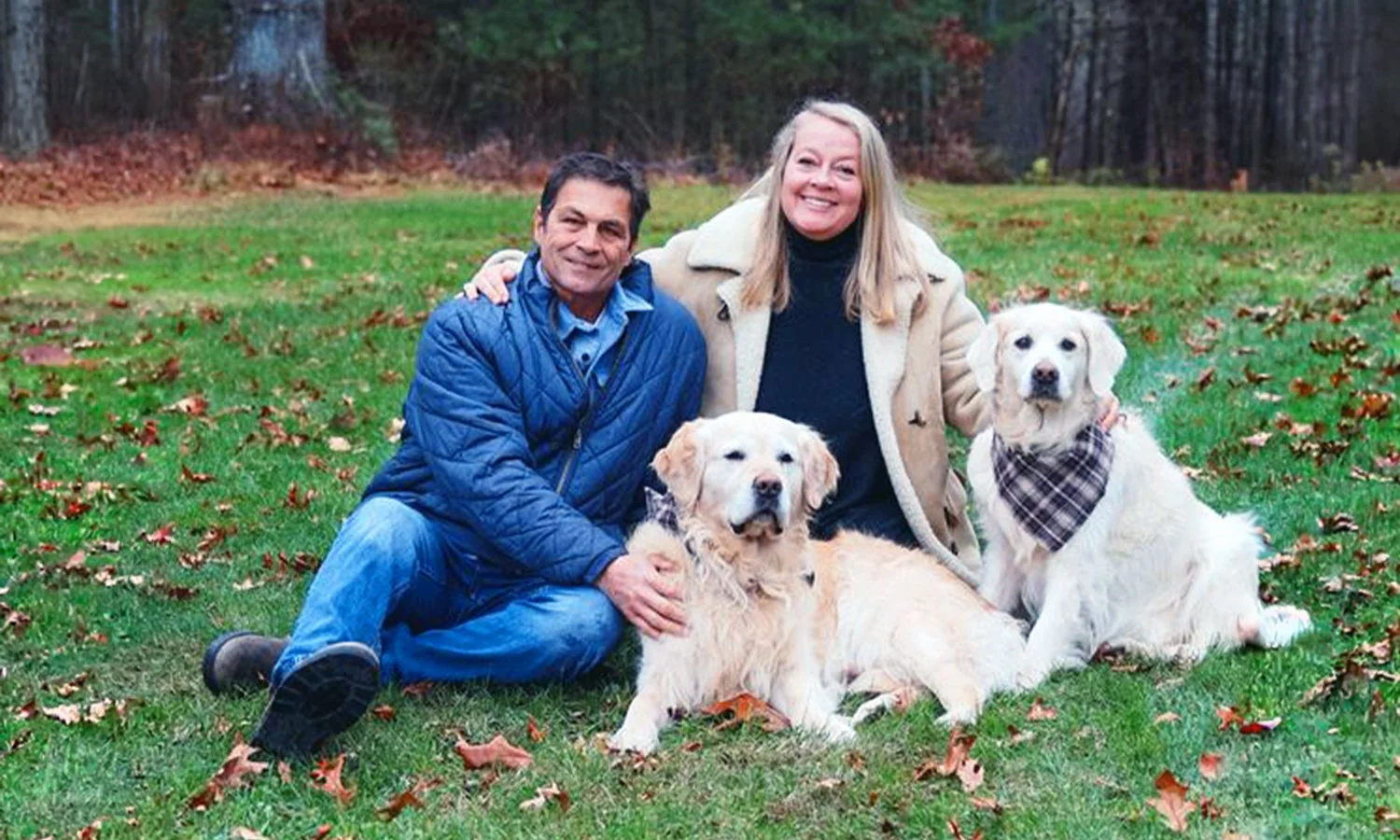 A man and woman posing outdoors on a grassy field with fallen autumn leaves, accompanied by two golden retriever dogs. The man and woman are sitting on the grass, her arm around one dog and the man, wearing a beige coat.