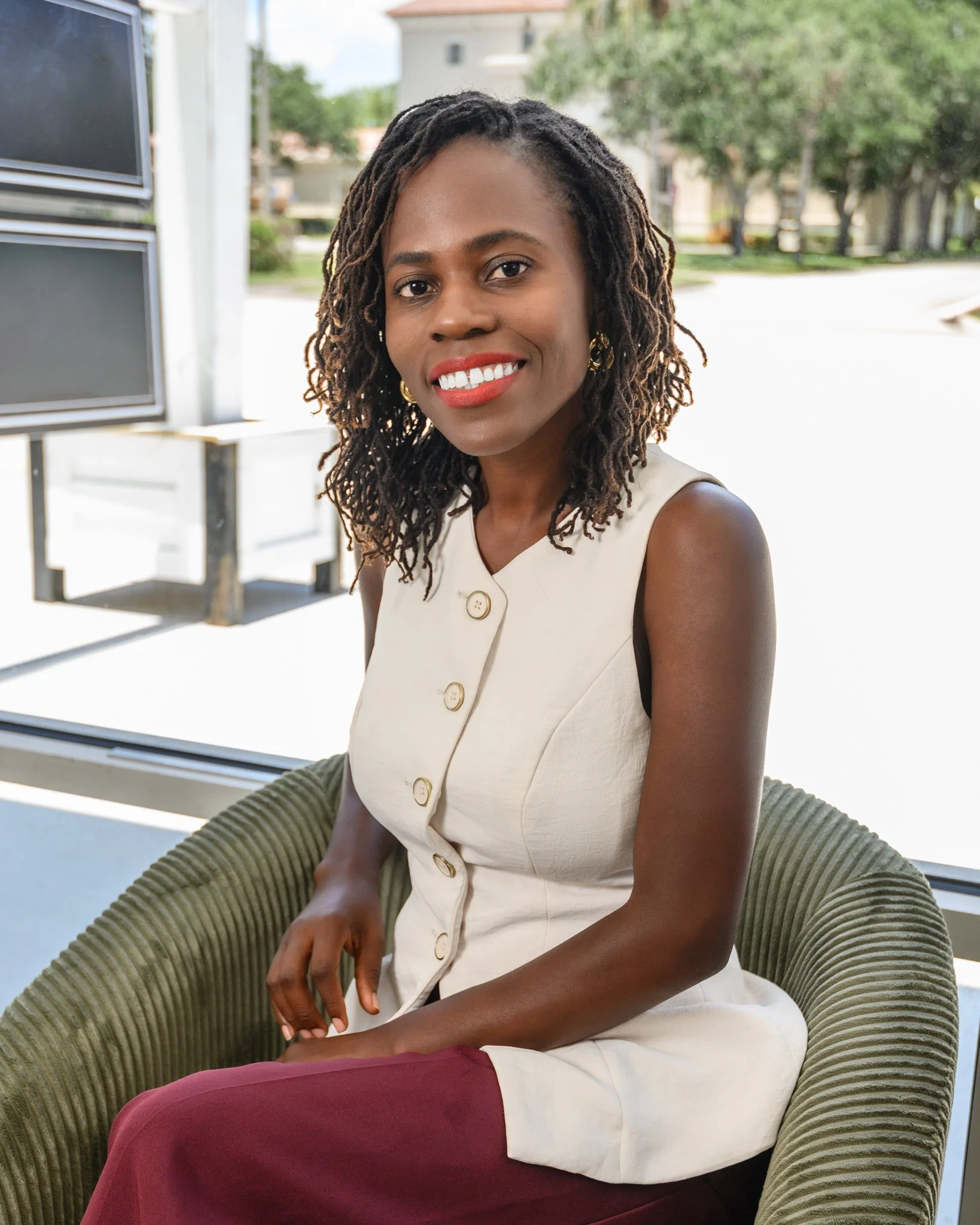 A woman with dark skin and curly dreadlocks sitting outdoors on a green striped chair, smiling at the camera. She is wearing a cream sleeveless dress with large buttons and red pants, with a blurred background of trees and buildings.