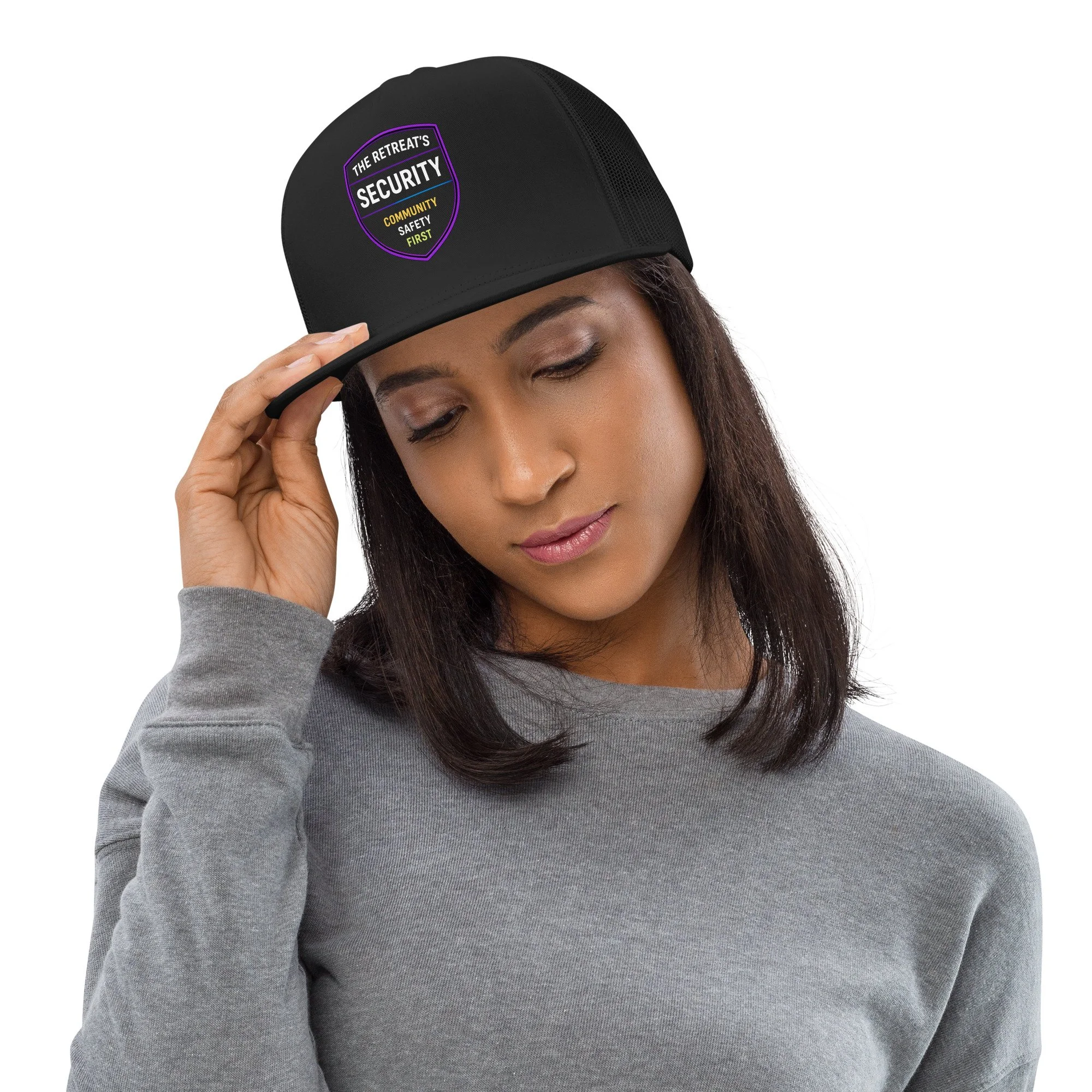 A woman with brown hair wearing a gray sweatshirt and adjusting a black baseball cap that has a security badge sticker on it.