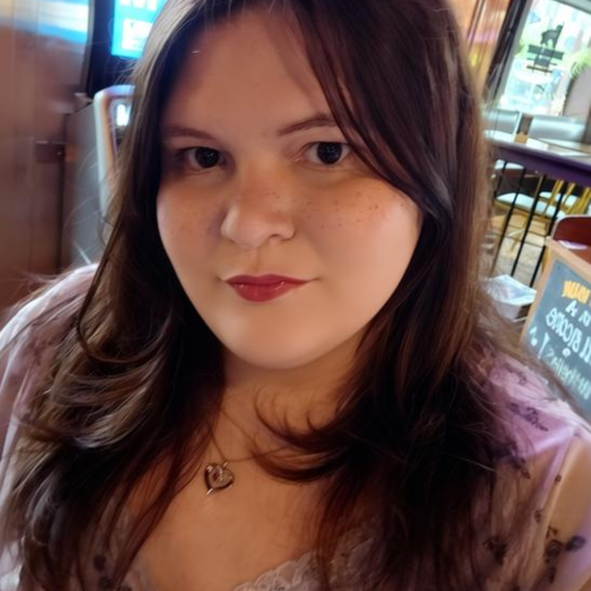 Close-up of a woman with long, wavy brown hair and dark eyes, wearing a ring and a heart-shaped necklace, inside a casual dining area.