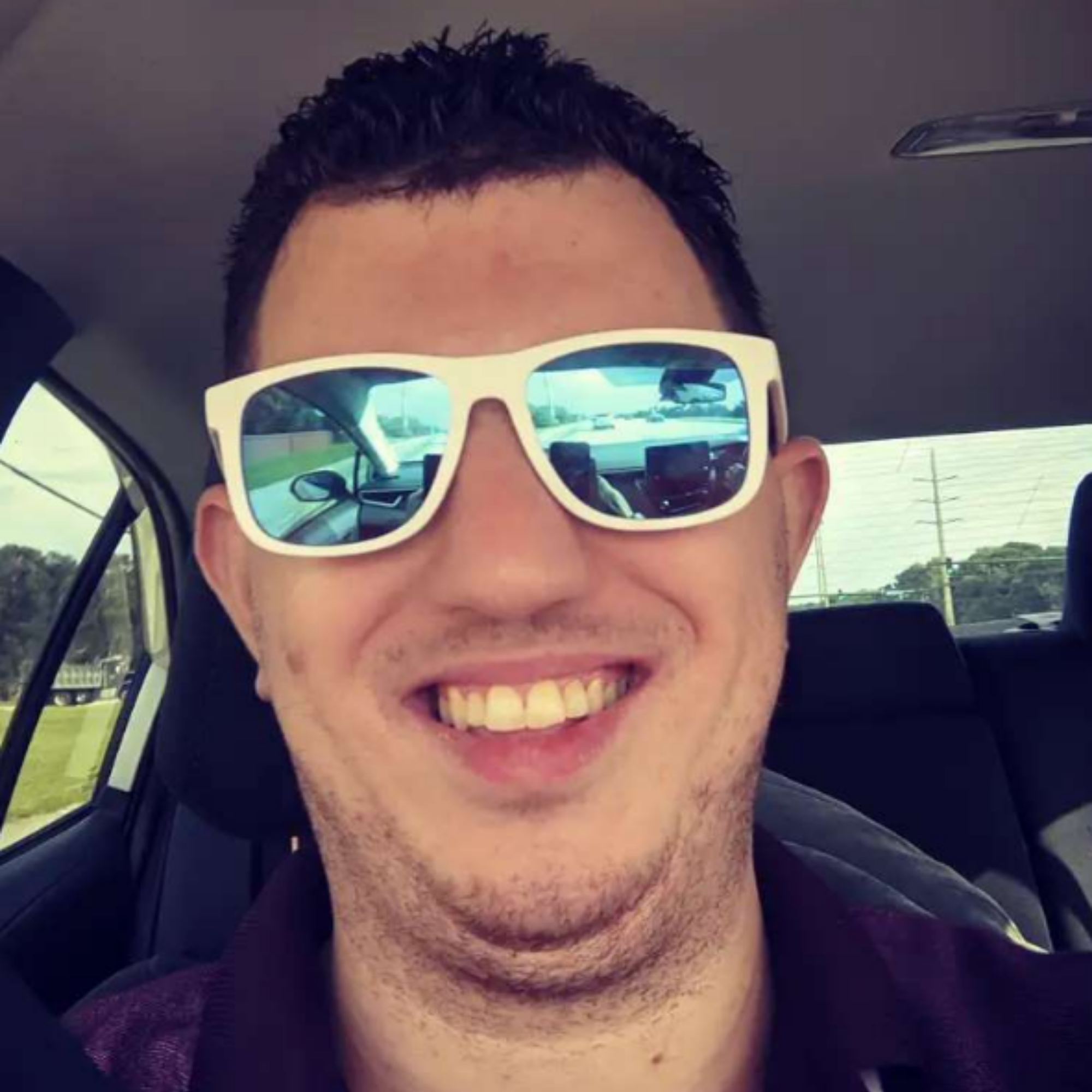 Close-up photo of a person with dark hair, wearing large white sunglasses, smiling, and showing their teeth.