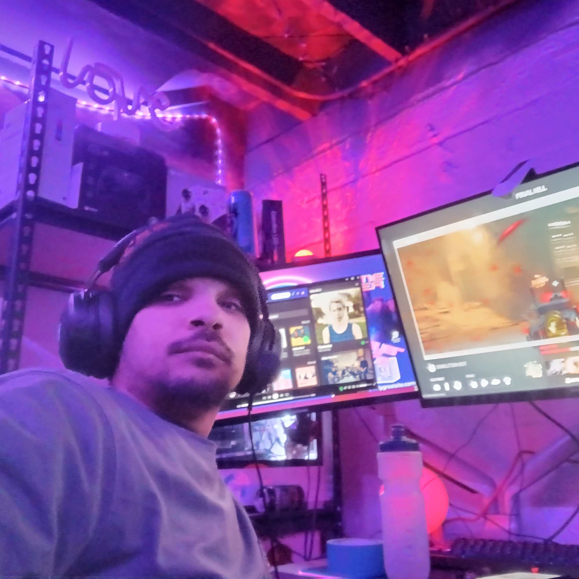 Person wearing gaming headphones in a room lit with purple and red neon lights, with monitors displaying colorful screens.