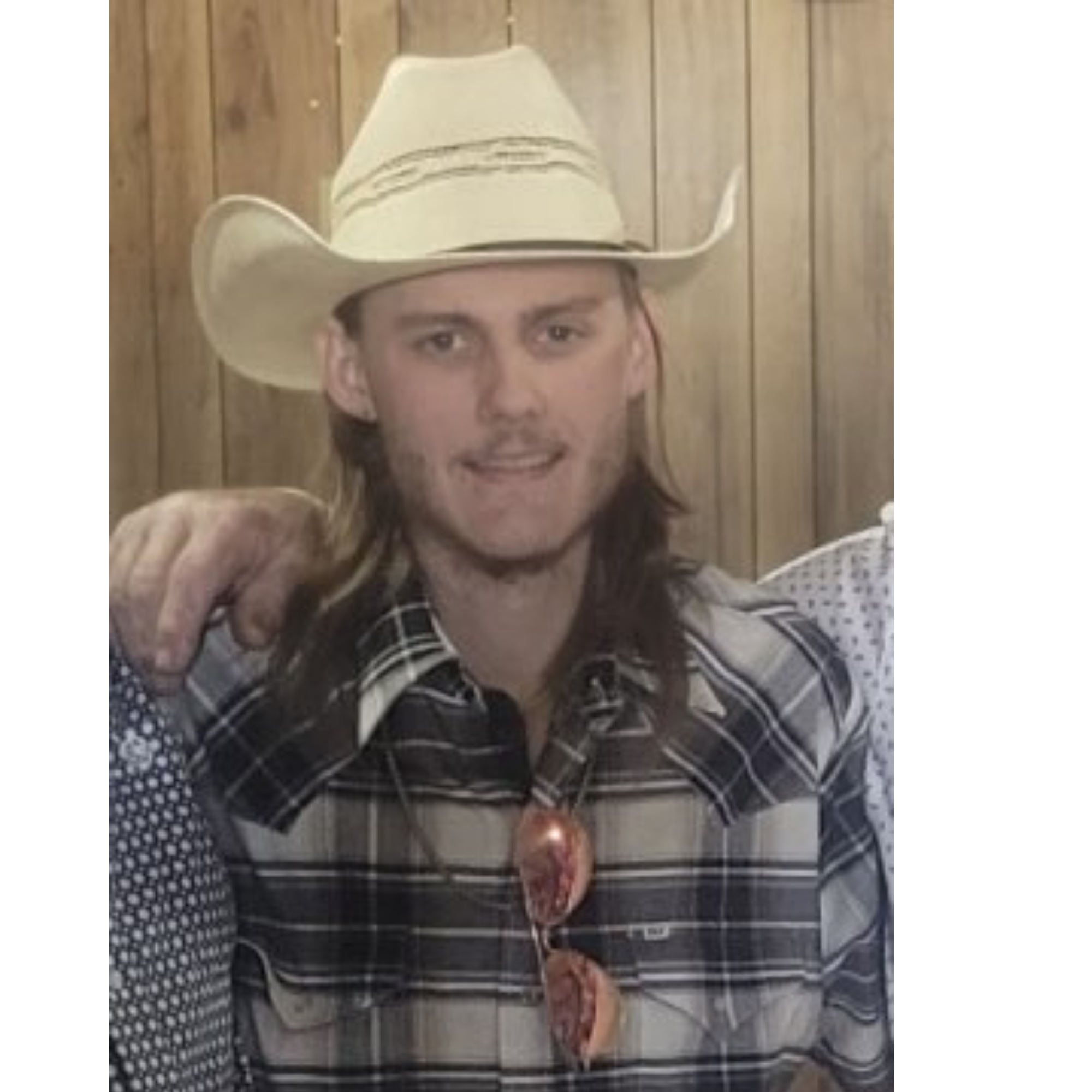 A person wearing a cowboy hat and a plaid shirt sitting beside a woman with long hair and a floral dress, with a wooden wall in the background.