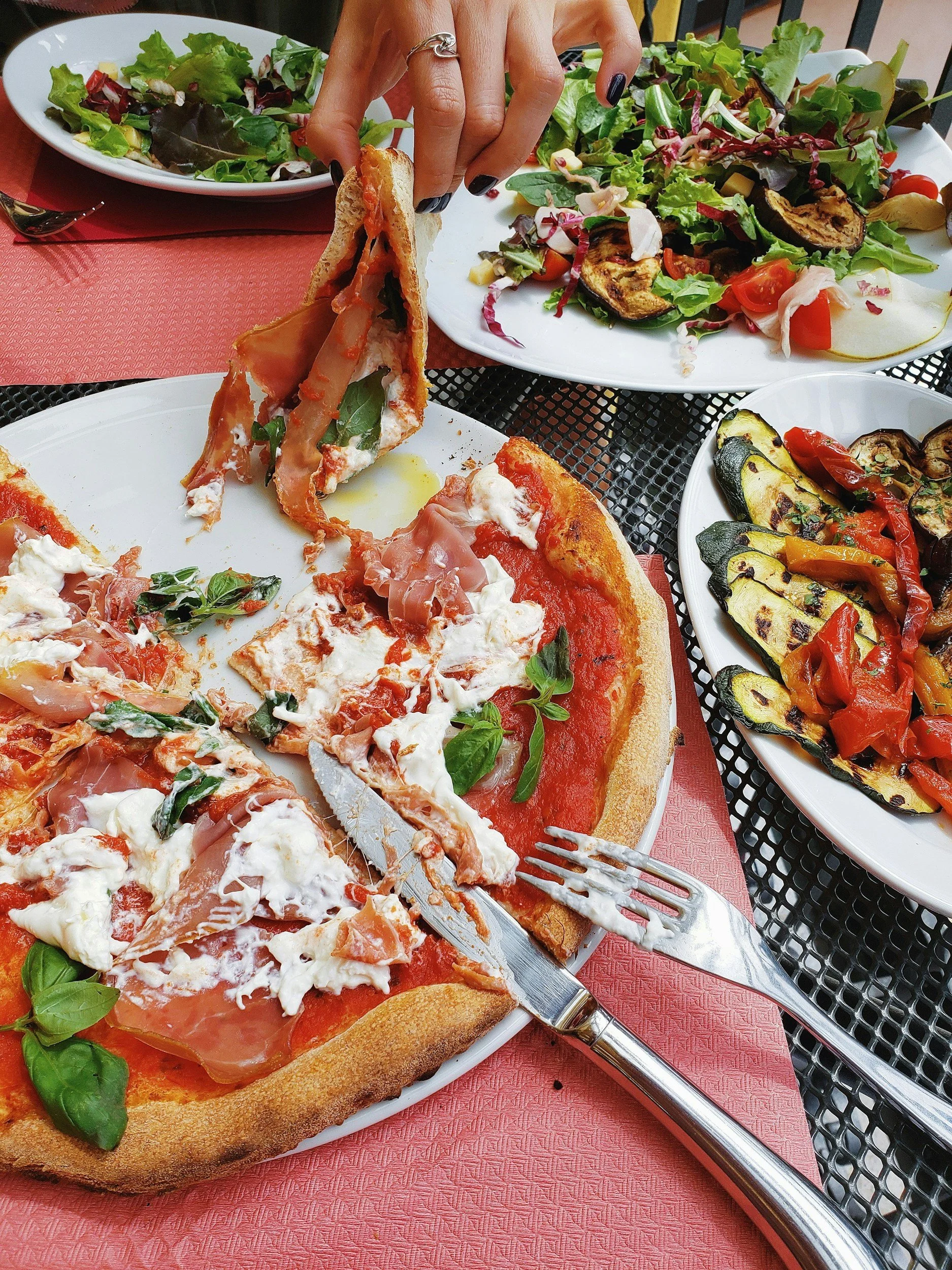 Eine Person isst eine Pizza mit Schinken, frischen Basilikumblättern, Tomatensauce und Käse, während andere Gerichte mit Salat und gegrilltem Gemüse auf Tischen mit roten Tischdecken sichtbar sind.