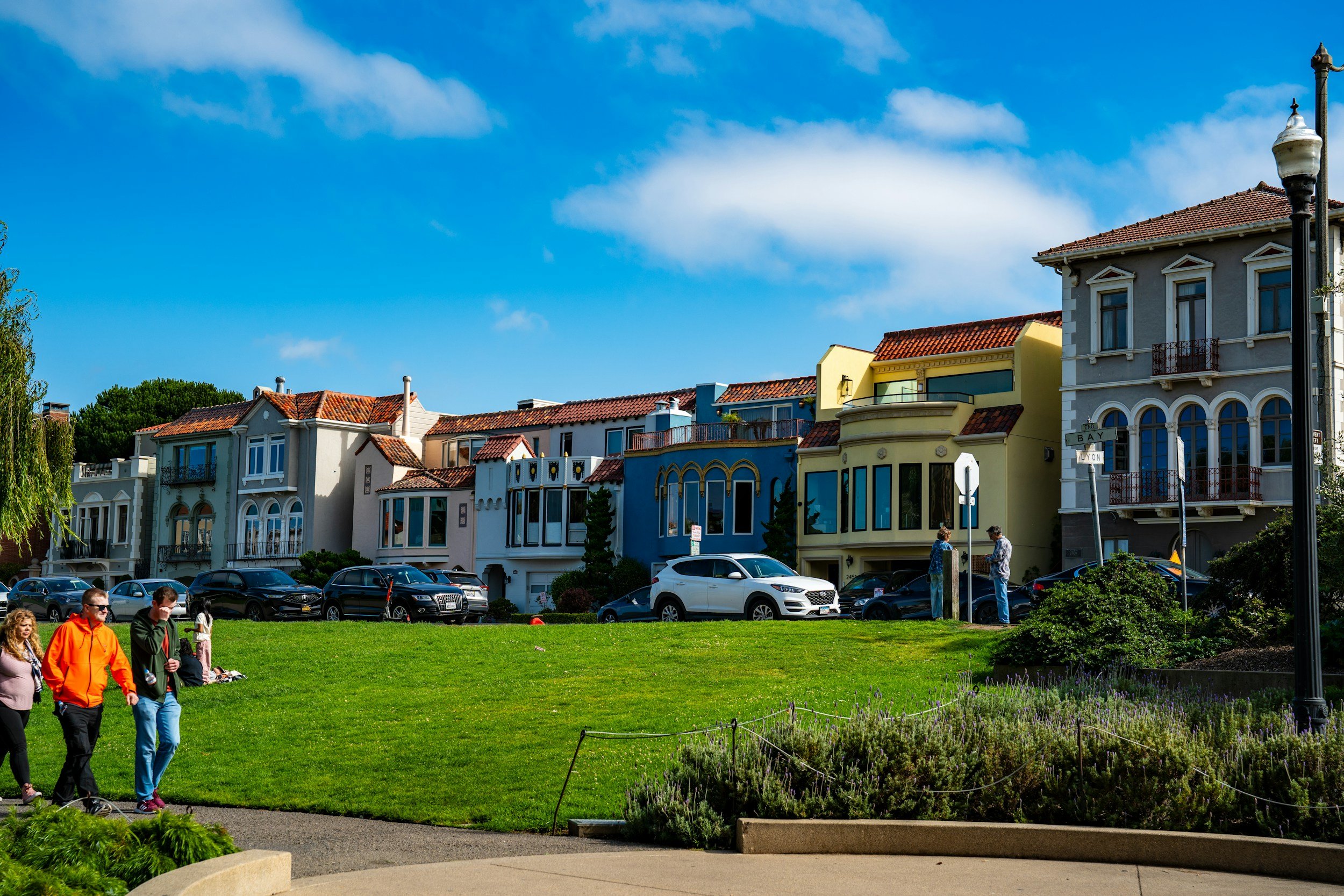 People walking and sitting on grass in a park with colorful houses and a blue sky with clouds in the background.