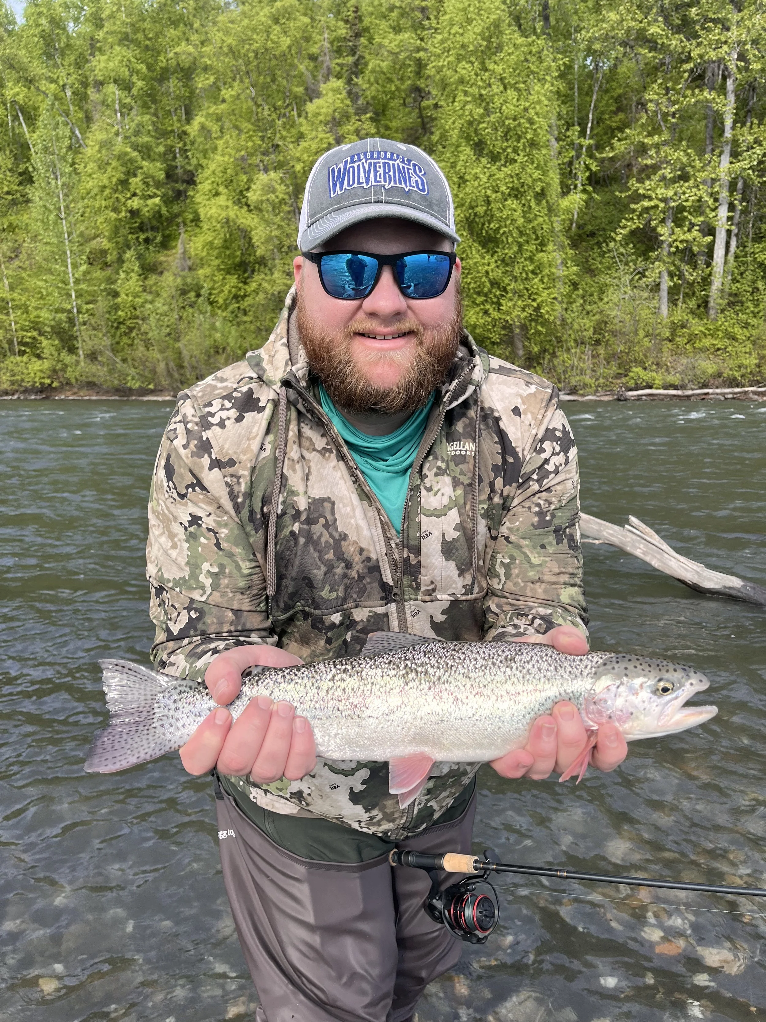 A man wearing sunglasses, a baseball cap with 'Wolverines' written on it, a camouflage jacket, and gray waders, holding a fish in a river with a forest in the background.