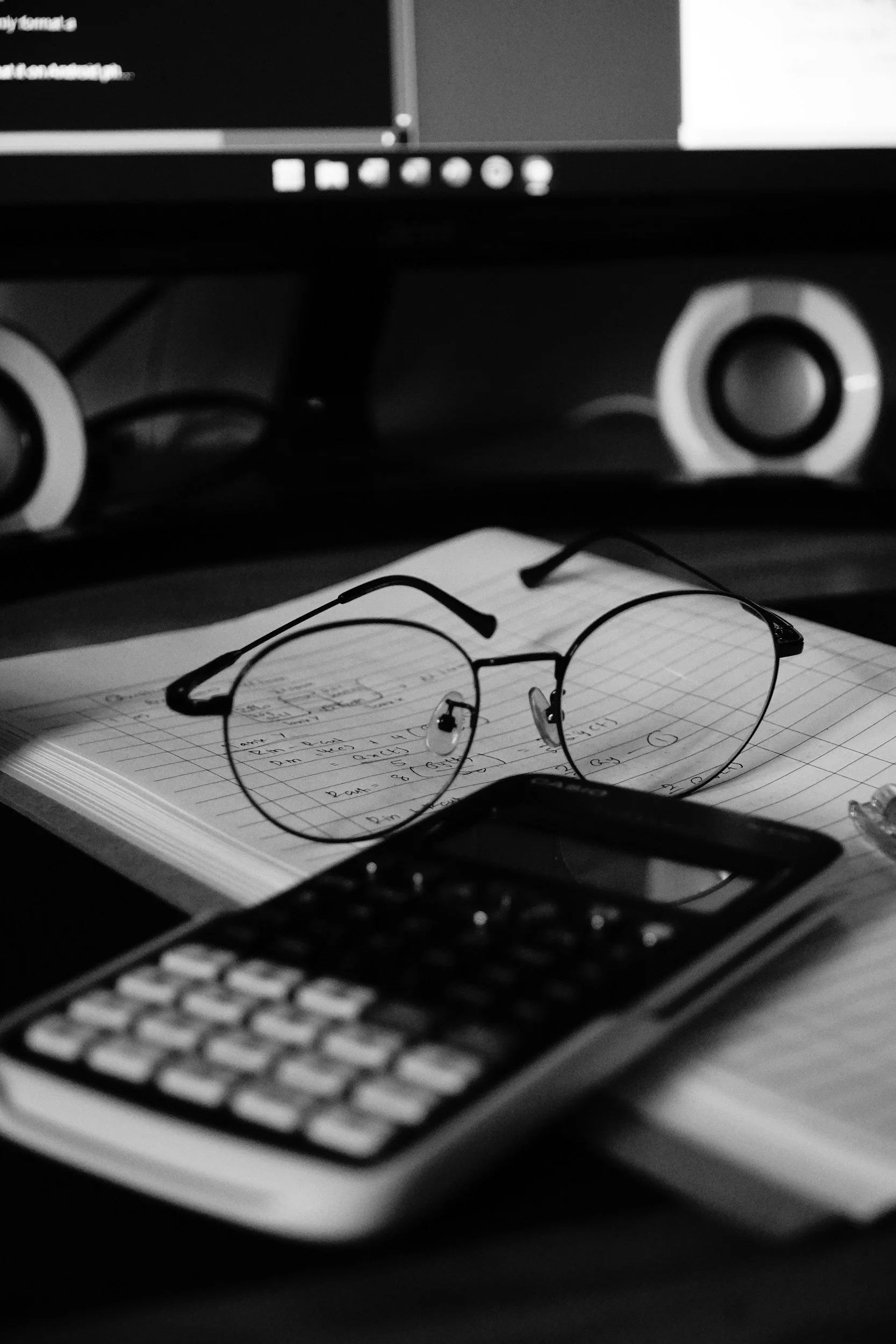 A pair of eyeglasses resting on a notebook with handwritten notes, with a keyboard and a computer monitor in the background.