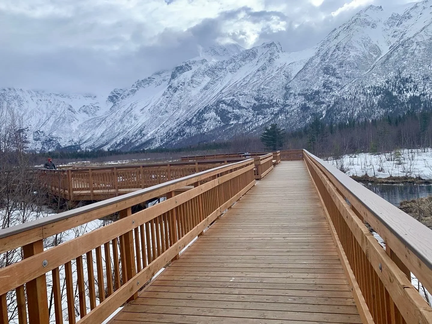 A wooden walkway over a river in a snowy, mountainous landscape with snow-covered peaks and cloudy sky in the background.