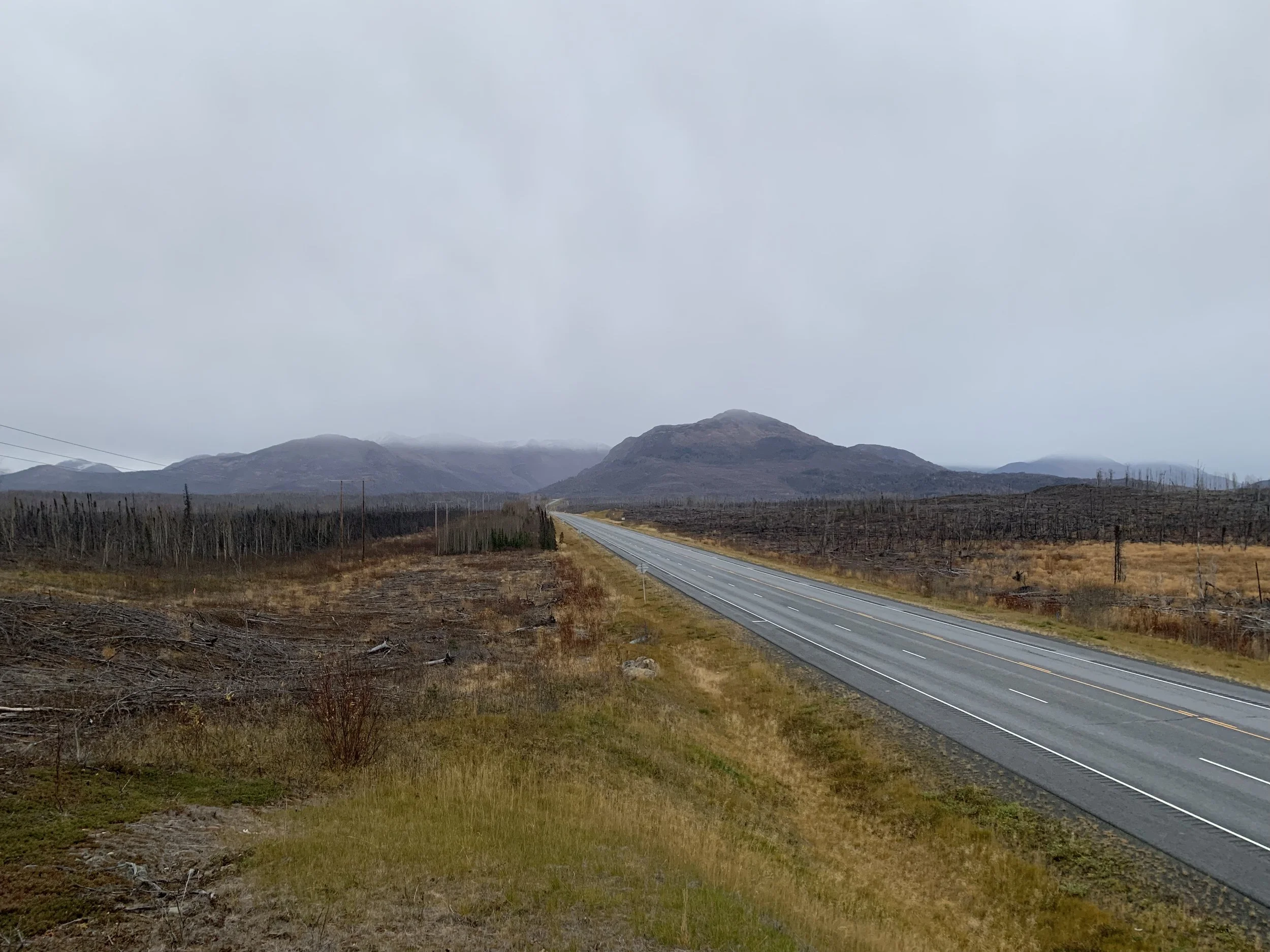 A long, straight highway cuts through a landscape of burned brown fields and low hills in a cloudy, overcast sky.