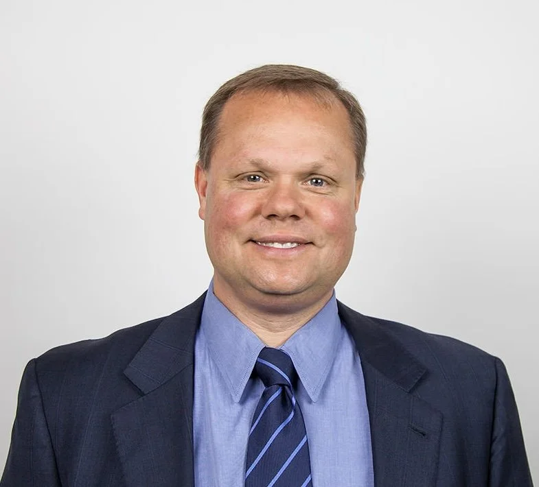 Headshot of a middle-aged man in a dark suit, light blue dress shirt, and a striped navy tie, smiling against a plain light gray background.