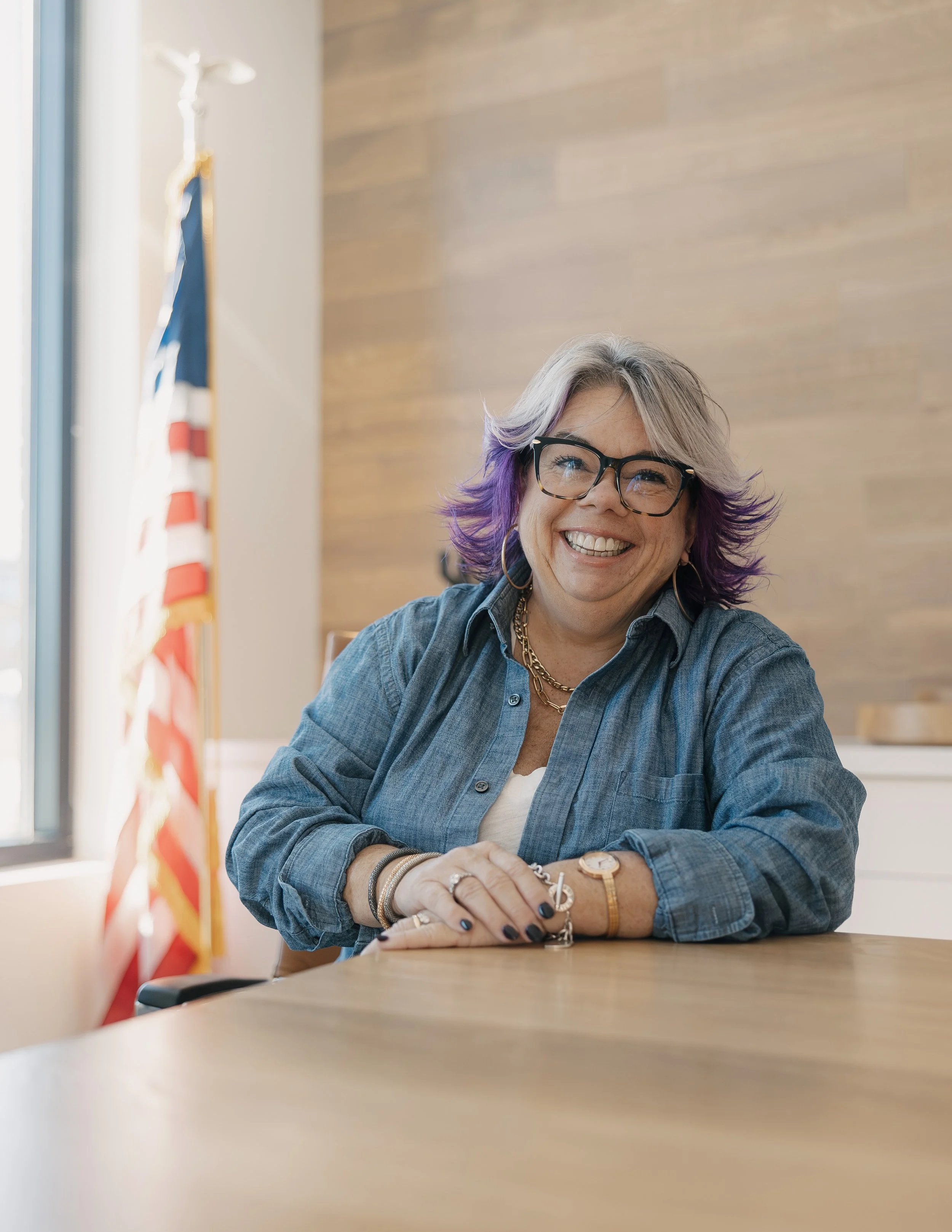 A woman with short gray and purple hair, wearing glasses and a denim shirt, smiling while sitting at a table in a room with wooden walls and an American flag in the background.