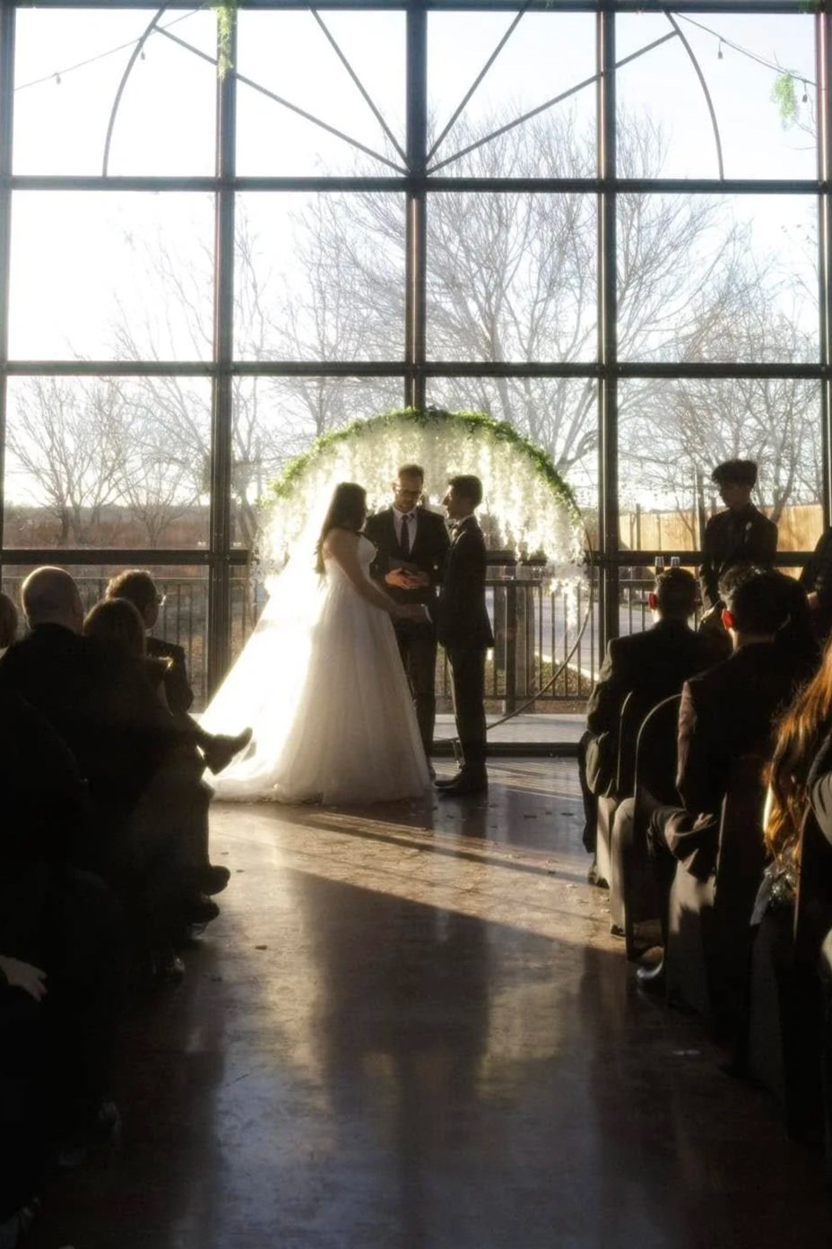 A wedding ceremony taking place indoors near large window with natural light, showing a bride and groom holding hands during their vows, with guests seated around them, and floral decorations behind them.