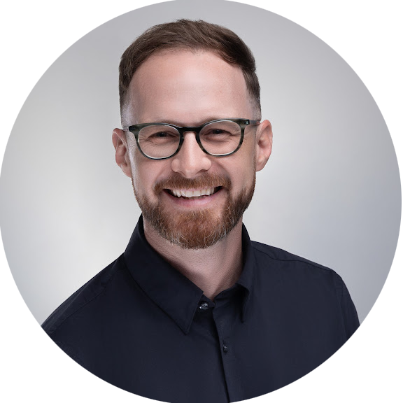 A man with glasses, a beard, and short hair smiling in front of a plain background.