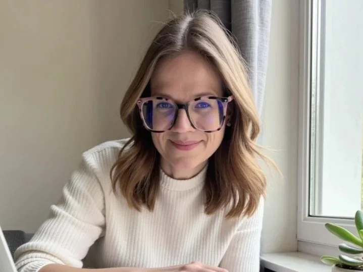 Dr Nicola Sutton, HCPC Registered Psychologist and founder of Sutton Psychology -- a woman with shoulder-length brown hair and glasses, smiling by a window with a small plant nearby
