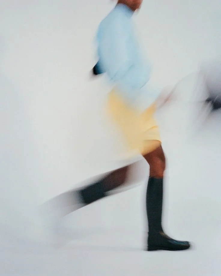 Blurred image of a person wearing yellow shorts, a light blue shirt, and black knee-high boots, walking or running against a plain background.