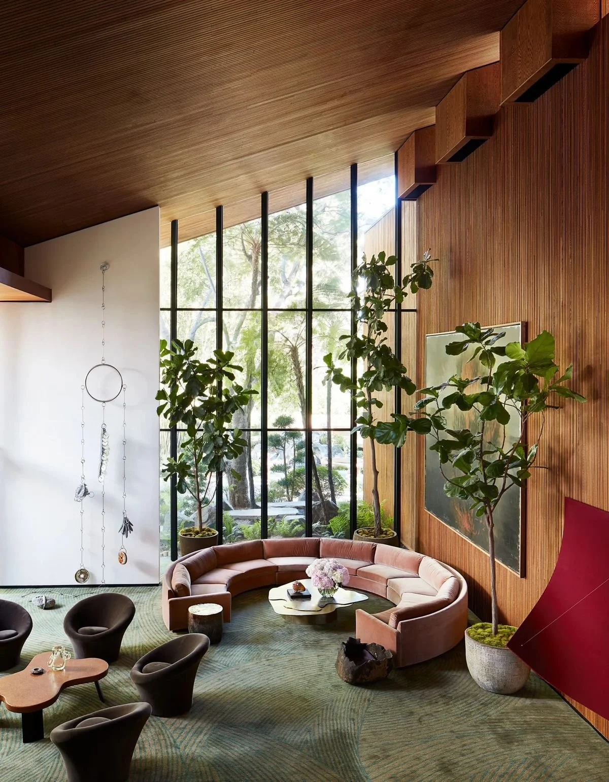 Interior of a modern living room with large floor-to-ceiling windows, a pink curved sofa, potted greenery, and wooden ceiling and walls.