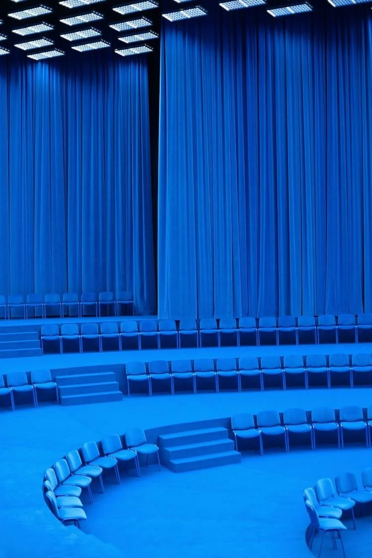 Empty theater with blue stage curtains and rows of blue chairs arranged in a circular manner.