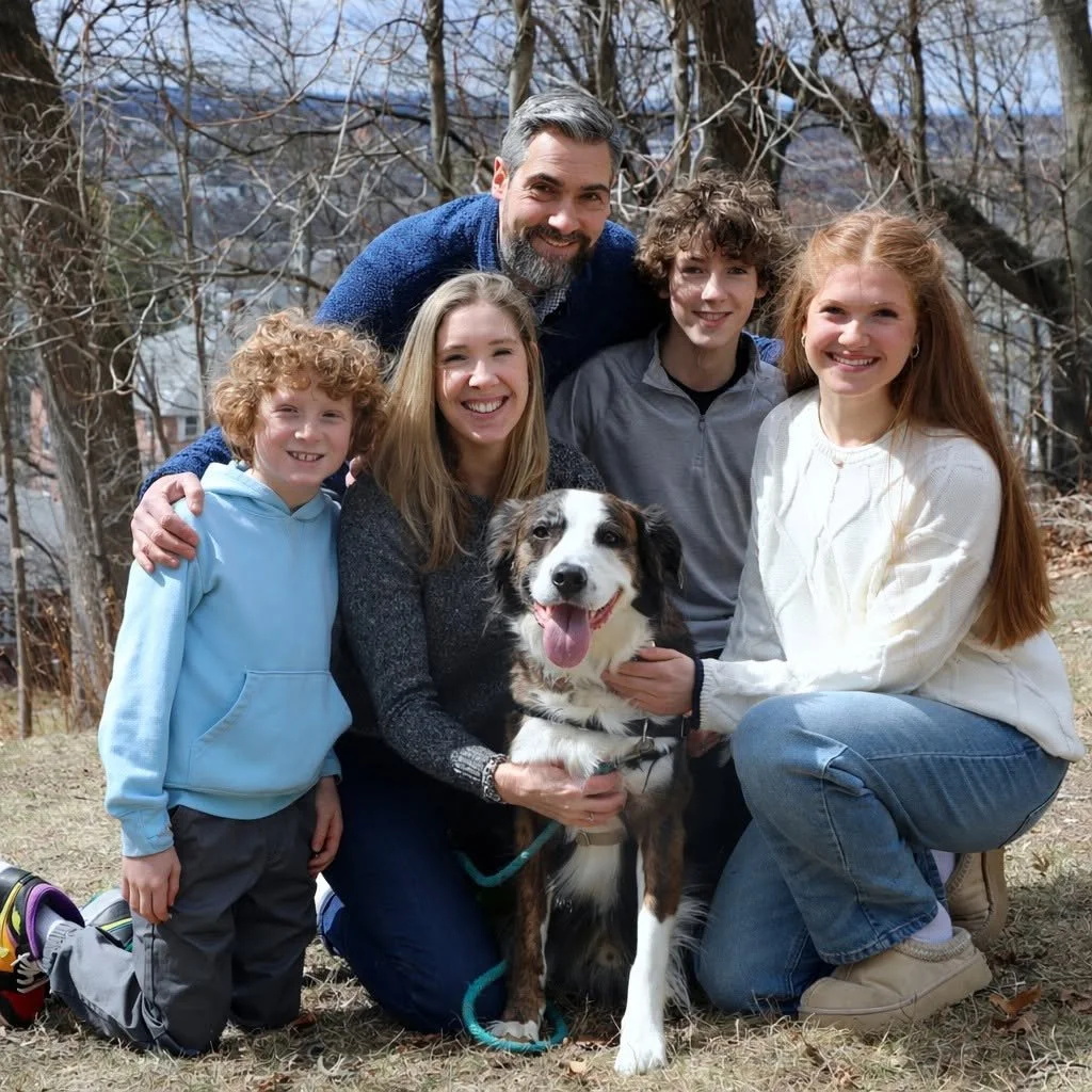 A family of six, including five people and a dog, outdoors on a sunny day with trees in the background, smiling and posing for a group photo.