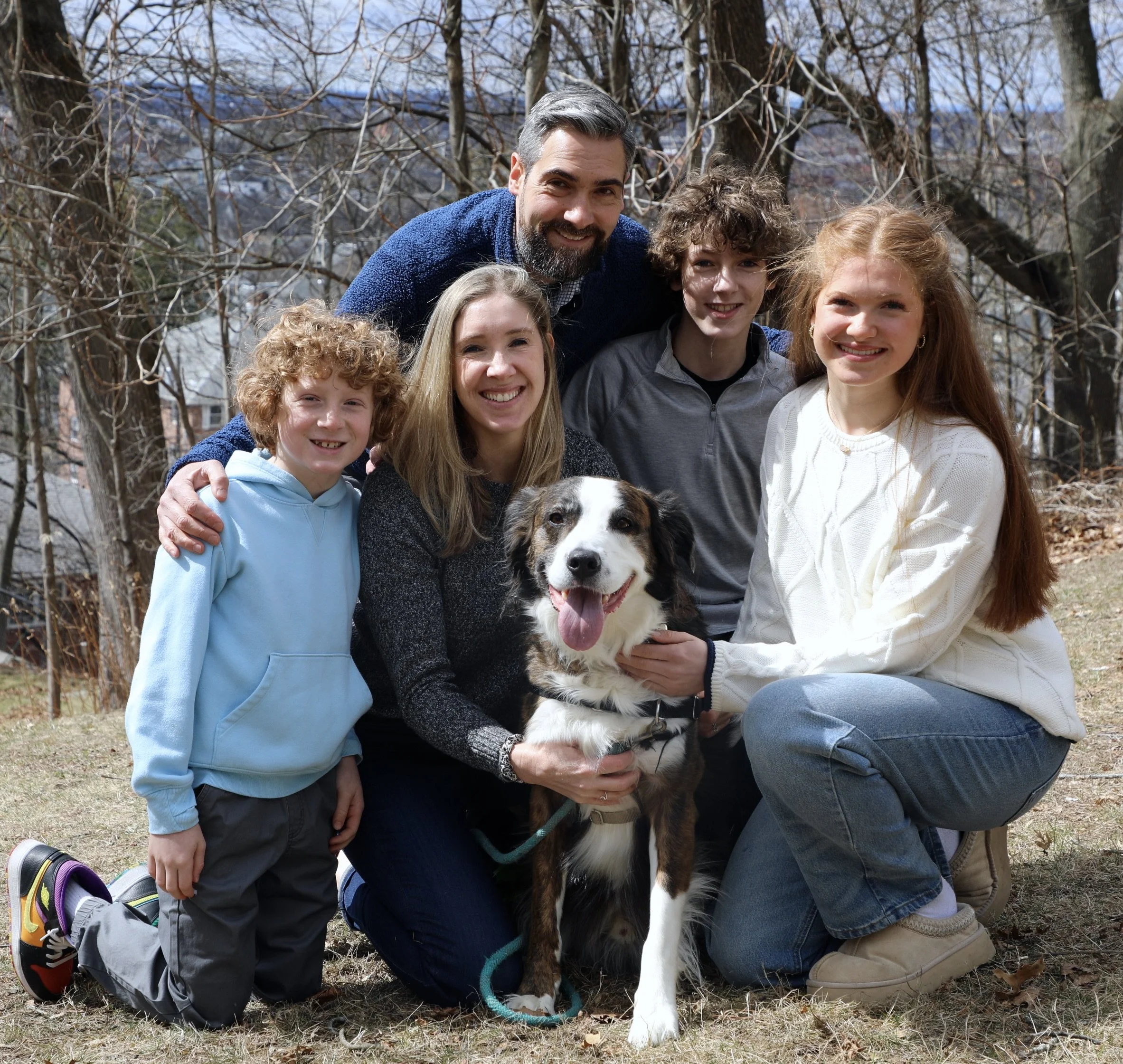 A group of five people and a dog outdoors on a sunny day, with trees and a cloudy sky in the background. They are smiling and posing for the photo.