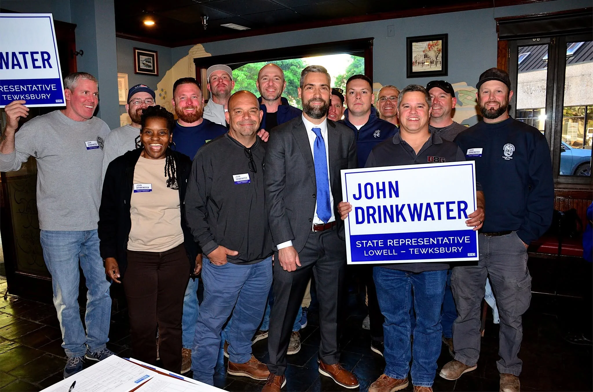 A group of people posing indoors, holding signs supporting a state representative candidate named John Drinkwater. The signs mention Lowell-Tewksbury. The group includes men and women of different ages, all smiling, with some wearing campaign shirts and badges.