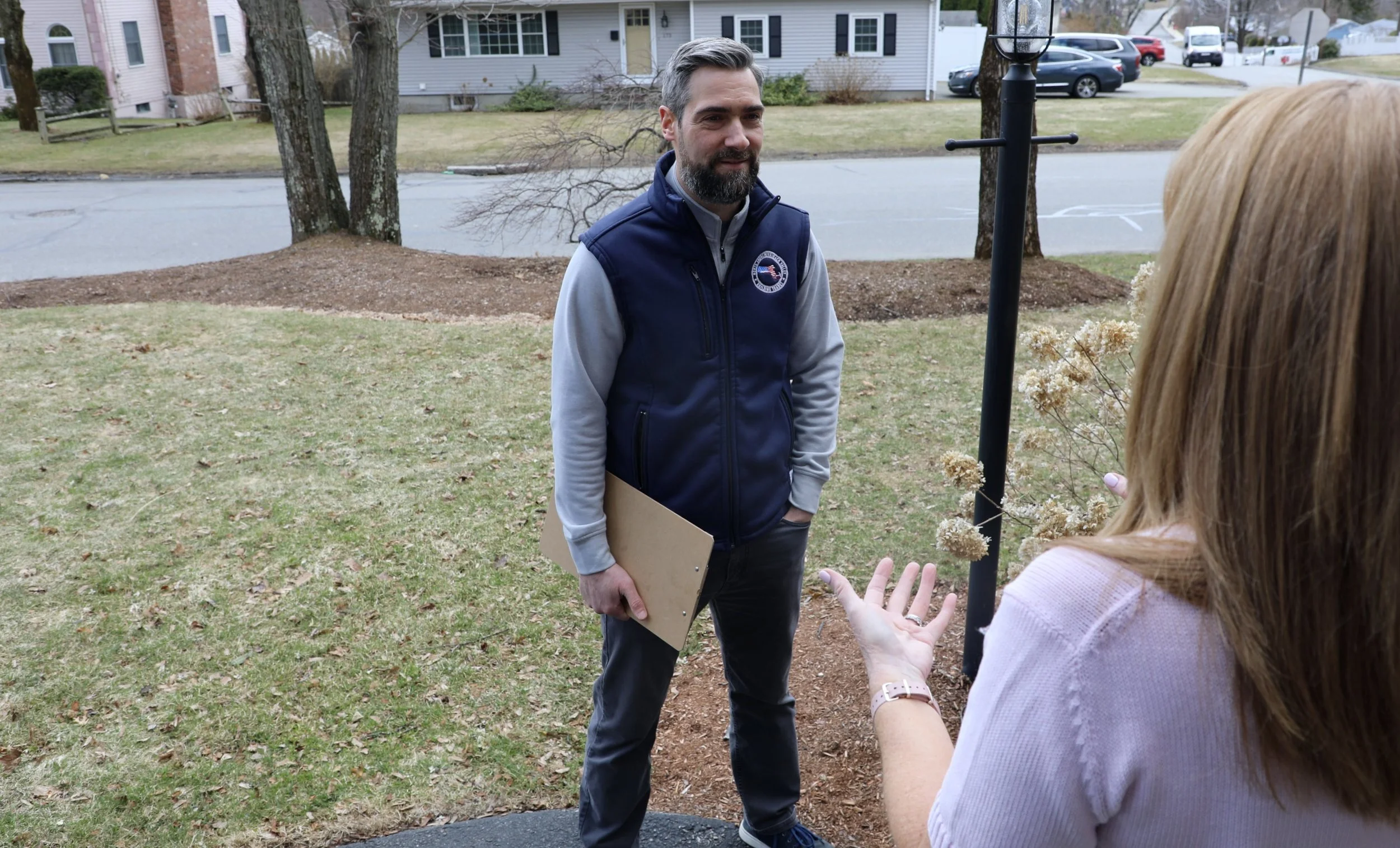 A man with gray hair and a beard wearing a navy vest with an emblem, talking to a woman with long brown hair, gesturing with her hands, outdoors in a grassy area with trees and houses in the background.