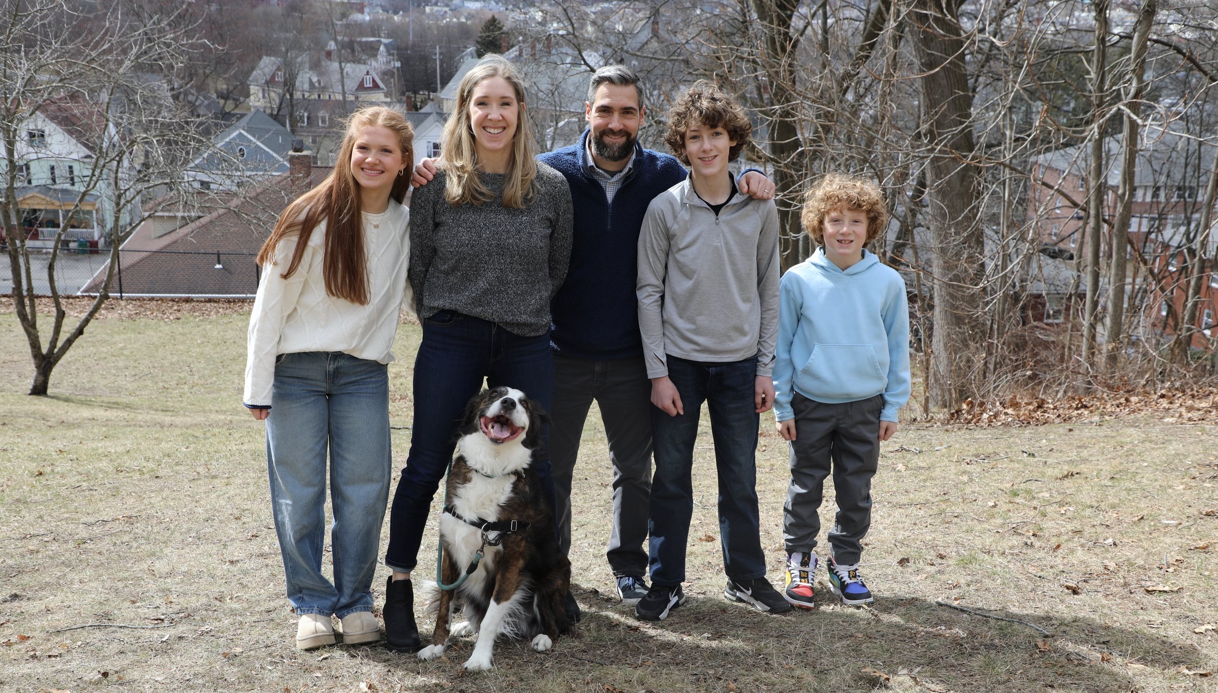 A family of five people and a dog standing outdoors in a park-like setting, with leafless trees and houses in the background, on a day with overcast weather.