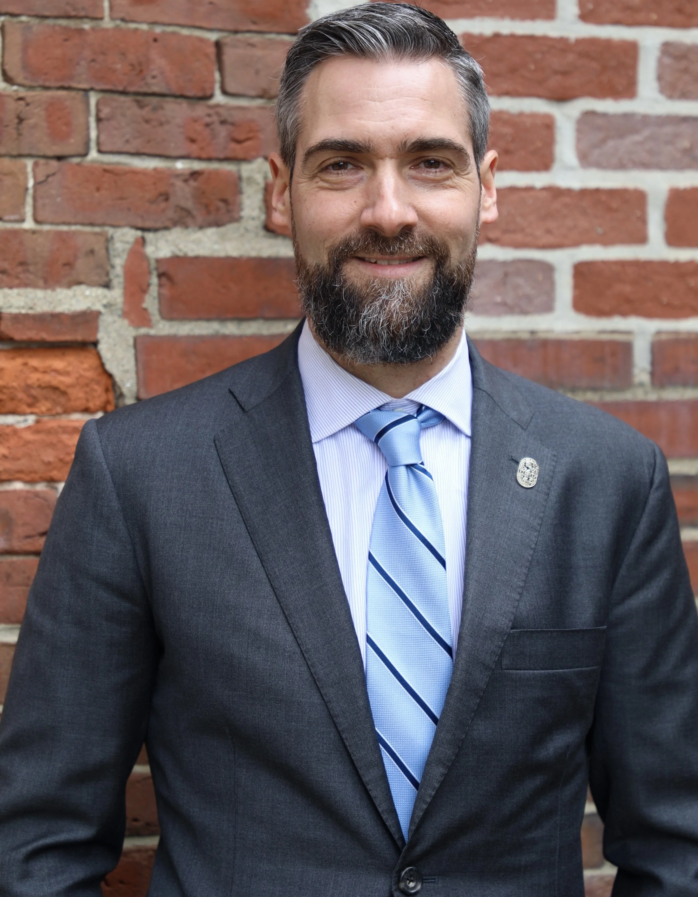 A man in a dark suit, light blue shirt, and light blue striped tie standing in front of a brick wall.