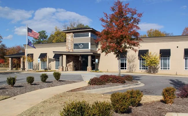 Tan, single-story GHFCU branch building with a stone-accented entrance. The exterior features dark awnings, a U.S. flag, and an autumn tree in the foreground.