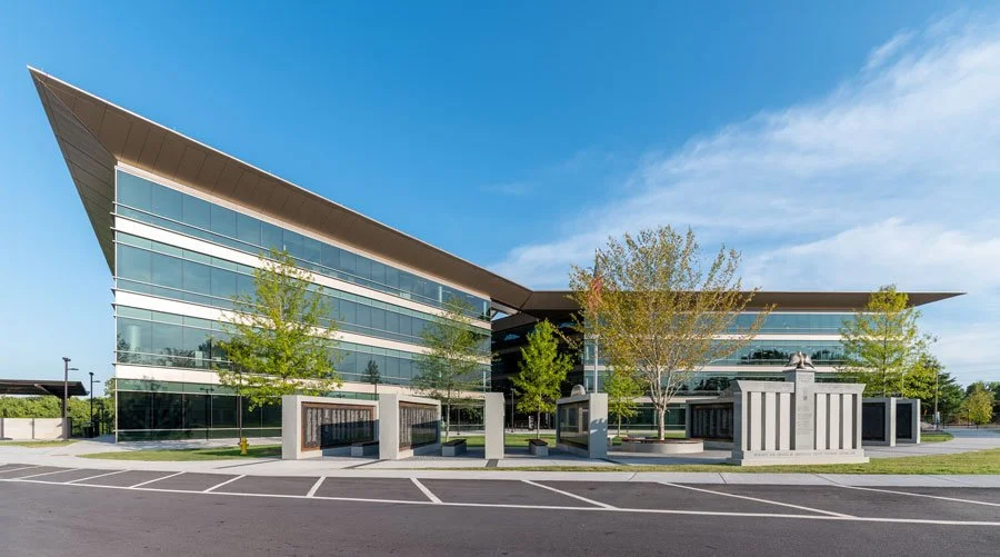 Modern, multi-story glass office building with a large angled roof overhang. A stone memorial plaza with plaques and an eagle statue sits in the foreground.